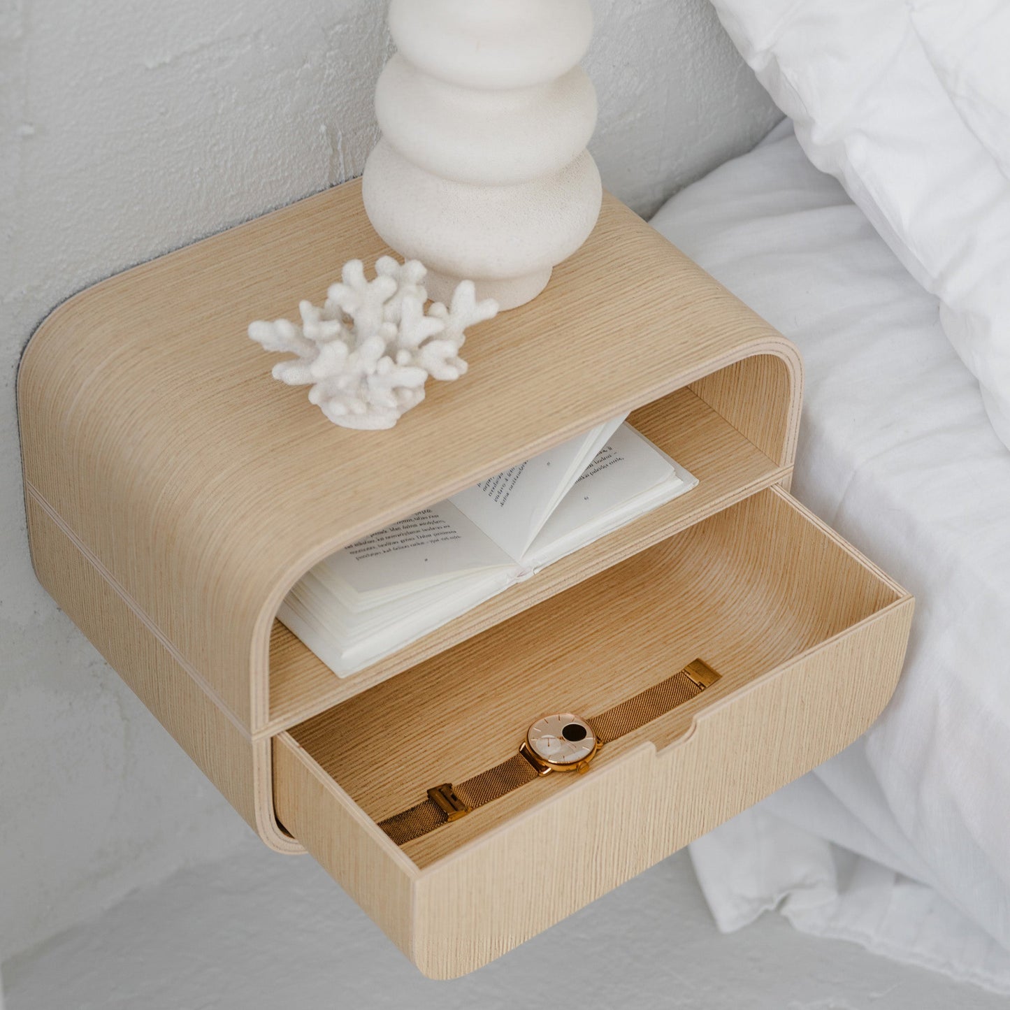 Minimalist wall-mounted oak nightstand with open drawer holding a watch and papers, styled in a room setting.