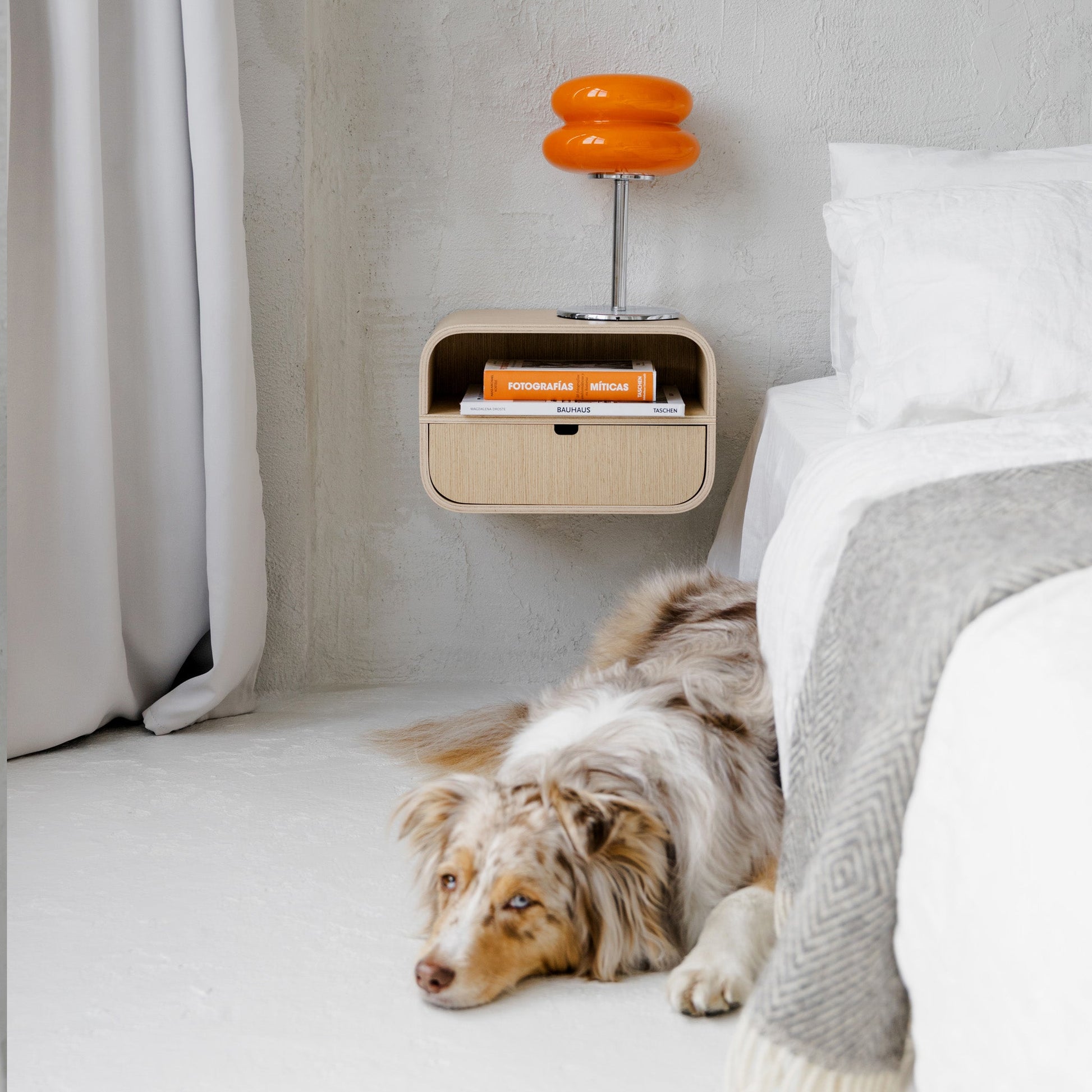 A modern wall-mounted nightstand with a single oak drawer and books on top, styled next to a bed with a dog lying nearby.